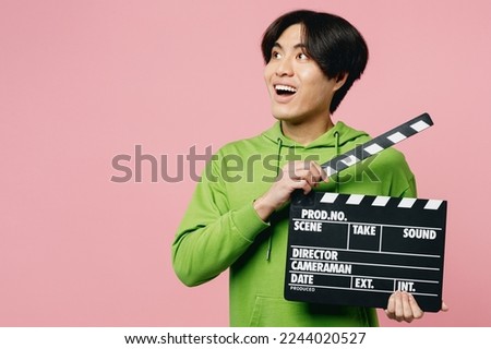 Young man of Asian ethnicity wear green hoody hold in hand classic black film making clapperboard look aside on workspace area isolated on plain pastel light pink background. People lifestyle concept