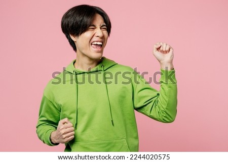 Young happy fun man of Asian ethnicity wear green hoody doing winner gesture celebrate clenching fists say yes isolated on plain pastel light pink background studio portrait. People lifestyle concept Royalty-Free Stock Photo #2244020575