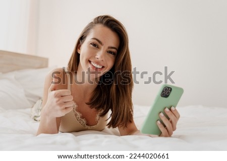 Happy young woman in casual casual clothes lying on her stomach hold in hand use mobile cell phone show thumb up like gesture rest relax spend time in bedroom lounge home in own room house good day