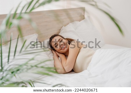 Smiling young woman in casual casual clothes lying in bed hands folded under head look camera rest relax spend time in bedroom lounge home in own room house wake up dream be lost in reverie good day Royalty-Free Stock Photo #2244020671