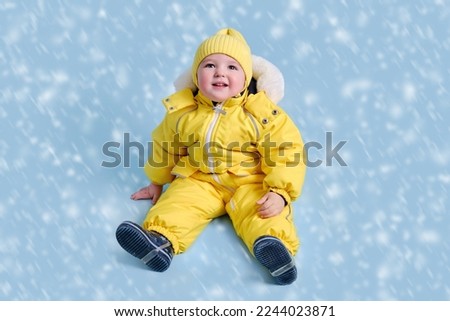 Happy toddler baby in winter clothes snowsuit on studio blue background. A child in a warm yellow jumpsuit with a hood. Kid aged one year five months Royalty-Free Stock Photo #2244023871
