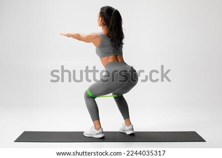 Athletic girl doing squats exercise for glute with resistance band on gray background. Fitness woman working out