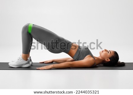 Athletic girl doing glute bridge exercise with resistance band on gray background. Fitness woman working out Royalty-Free Stock Photo #2244035321
