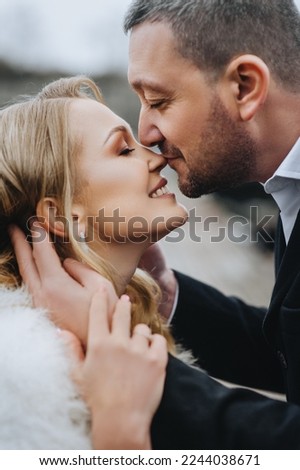 Stylish bearded groom in a black coat and a beautiful smiling blonde bride in a white dress, fur coat hugging, gently kissing outdoors in autumn. Wedding photography, close-up portrait.