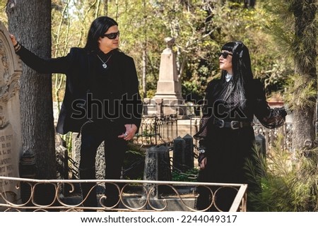Stylish dark gothic couple with black clothes between graves in cemetery in a sunny day