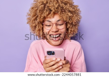 Positive curly haired young woman smiles happily and focused at smartphone screen reads funny story dressed in casual pink pullover wears transparent eyeglasses isolated over purple background. Royalty-Free Stock Photo #2244057881