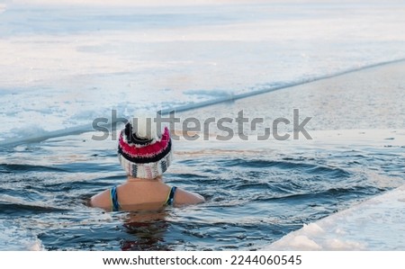 A woman in a swimsuit in a warm hat swims in an ice hole in winter swimming, winter therapy, ice swimming in the river hardening of the body. Royalty-Free Stock Photo #2244060545