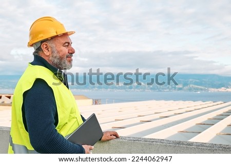 Portrait of smiling middle aged bearded supervisor in hardhat and safety vest with tablet on building site. Structural engineer or architect monitors the progress of the work on construction site.