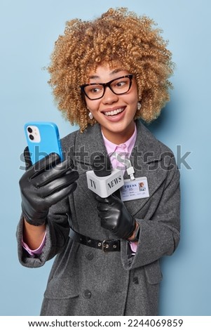 Happy smiling woman correspondent holds microphone smiles broadly uses smartphone for recording video makes broadcasting dressed in elegant clothes isolated over blue background. Press concept Royalty-Free Stock Photo #2244069859