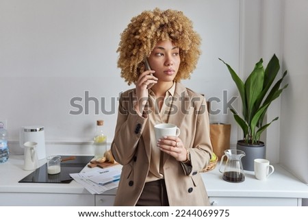 Indoor shot of pensive beautiful curly haired woman dressed elegantly has telephone conversation and drinks coffee poses at home kitchen talks with business partner thinks over future plans.