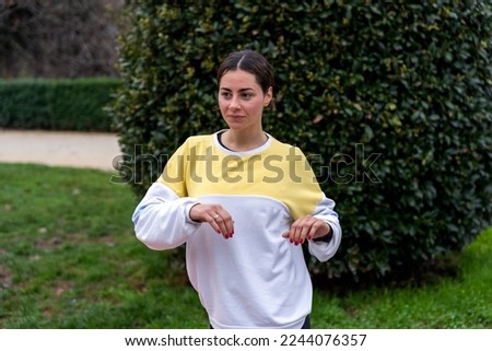 Positive young female in colorful sweatshirt with bun hairstyle standing near green bushes and doing warm up exercises in park