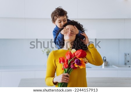 Playful hispanic curly boy stands on kitchen table puts palms on mother's eyes from behind dares flowers on mothers day. Curly Italian young woman gets colourful tulips from son. Cute family moments. Royalty-Free Stock Photo #2244157913