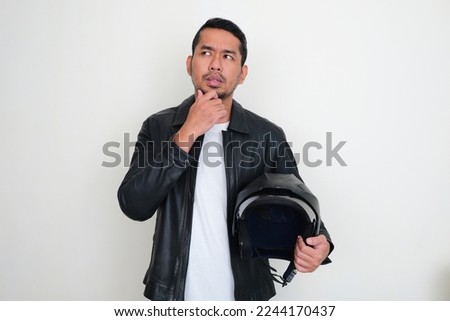Adult Asian man wearing leather jacket holding motorcycle helmet showing thinking expression Royalty-Free Stock Photo #2244170437