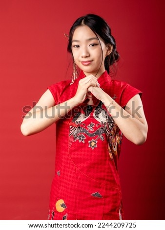 A 20-year-old Chinese Indonesian (Asian) girl in a Chinese traditional red dress (Cheongsam), with her hand, folded over each other as a gesture to greet Happy CNY. Isolated on a red background. Royalty-Free Stock Photo #2244209725