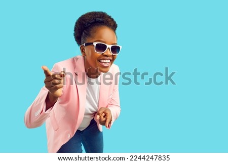 Happy young fashion model isolated on blue copy space background. Confident attractive African American woman in modern stylish pink jacket and trendy glasses smiles, looks at camera and points at you