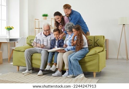 Friendly happy smiling big family with parents, kids, grandparents looking at family photo album together sitting in modern light living room at home. Emotional family moments, togetherness concept.