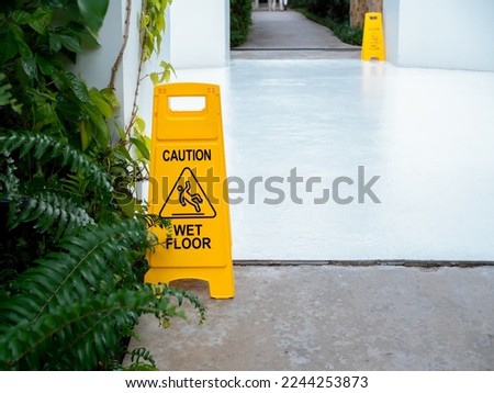 Yellow caution slippery wet floor sign with slippery person warning icon on wet white concrete floor background in front of the building near green leaves garden after raining with copy space.