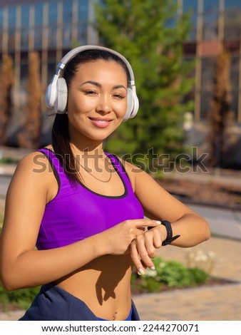 Vertical photo of a young asian woman checks her pulse during the race, smiling and looking at camera. The concept of modern technologies in sports.