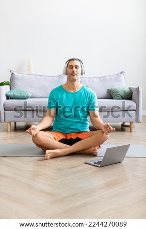Photo of a man meditates at home - using from the internet to monitor the workout. He listens to relaxing meditation music through headphones. Home workout and healthy lifestyle concept.