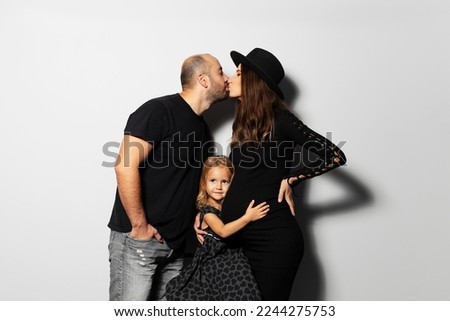 Studio portrait of young happy family, mother and father kissing, daughter hugging belly, on white background. Wearing hat, dressed in black.