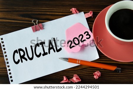 CRUCIAL 2023 - words on a white sheet on a wooden brown background with a cup of coffee and a pen