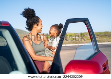 Cheerful mother embracing her cute little girl and looking at each other standing outside red car. Black female kid in mother's arms laughing near the car during travel. African family enjoying car. Royalty-Free Stock Photo #2244302309