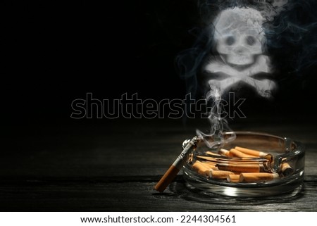 No Smoking. Skull and crossbones symbol of smoke over ashtray with stubs and smoldering cigarette on dark wooden table against black background. Space for text
