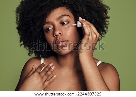 African American female with curly hair dropping moisturizing serum on cheek and looking away during skin care routine against green background Royalty-Free Stock Photo #2244307325