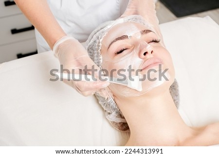 Beautician applies vitamins cream mask on woman face for rehydrate face skin, anti aging cosmetic procedure in beauty spa salon. Cosmetologist in gloves holds cosmetic brush for applying mask Royalty-Free Stock Photo #2244313991