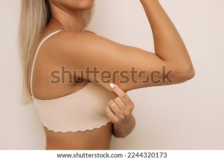 Cropped shot of a young blonde woman grabbing skin on her upper arm with excess fat isolated on a light beige background. Pinching the loose and saggy muscles. Overweight concept Royalty-Free Stock Photo #2244320173