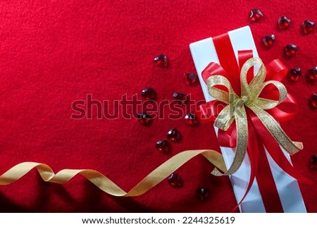 White gift box with red and gold ribbon, and a small red hearts shape on a red velvet floor with copy space, flat lay, Valentine's day background.