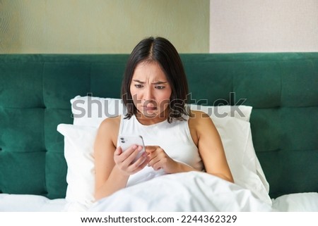 Bedroom picture of asian woman lying in bed, looking scared and shocked at smartphone screen, reading message with concerned face.