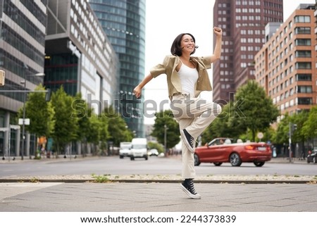 Portrait of asian happy girl jumping and dancing in city centre, posing on streets, express joy and excitement. Copy space Royalty-Free Stock Photo #2244373839