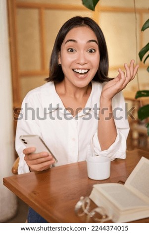Portrait of asian girl looking surprised, holding mobile phone with amazed face, great news on smartphone, sitting in cafe with book.