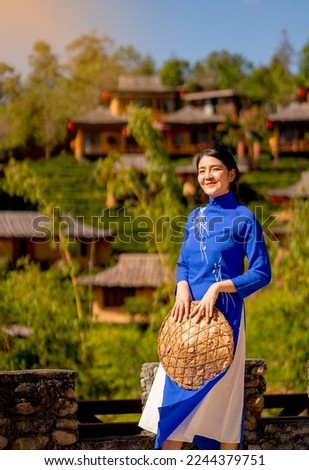 Vertical image of Asian girl hold hat and blue dress look like vietnamese style and stay in front of layering house village on mountain as background and look happy.  