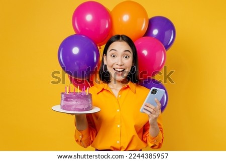 Happy fun surprised young woman wearing casual clothes celebrating near balloons hold cake with candles use mobile cell phone isolated on plain yellow background. Birthday 8 14 holiday party concept