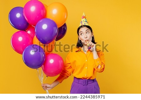 Happy fun young woman wear casual clothes hat celebrating hold look at bunch of colorful air balloons cake with candle blow pipe isolated on plain yellow background Birthday 8 14 holiday party concept