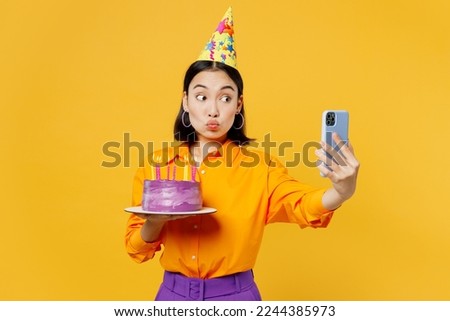 Happy fun young woman wearing casual clothes cap hat celebrating hold cake with candles using mobile cell phone doing selfie isolated on plain yellow background. Birthday 8 14 holiday party concept