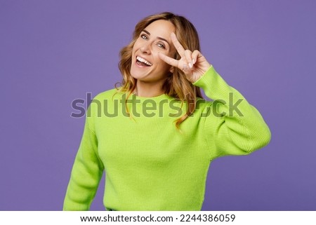 Young fun woman 30s she wearing casual green knitted sweater covering eye with v-sign victory gesture look camera isolated on plain pastel purple background studio portrait. People lifestyle concept