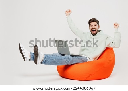 Full body young overjoyed happy fun cool IT man wear mint hoody sit in bag chair hold use work on laptop pc computer do winner gesture isolated on plain solid white background People lifestyle concept