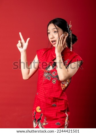 A 20-year-old Chinese Indonesian (Asian) girl in a Chinese traditional red dress (Cheongsam or Qipao), looks surprised as she is pointing to the copy space. Isolated on a red background.