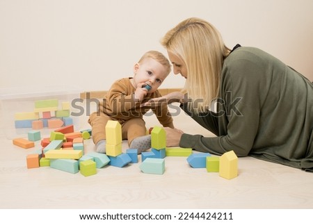 Mom plays with baby in the room,  child put wooden block in his mouth. Mom takes care of her son. Concept of time together, early development, safe toys, danger of small child to choke. Indoor.  Royalty-Free Stock Photo #2244424211