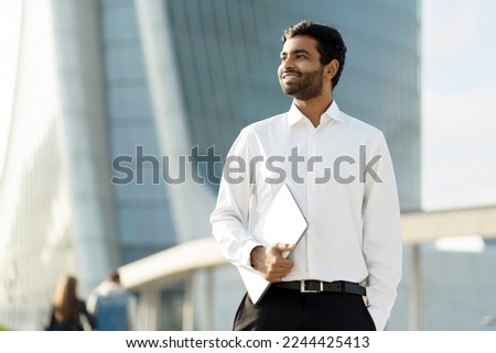 Confident smiling Indian businessman wearing white shirt holding laptop looking away on urban street. Successful business Royalty-Free Stock Photo #2244425413