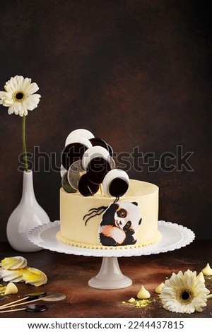 Baby birthday cake. Delicate pastel yellow festive cake decorated with lollipops and panda picture over dark brown concrete background. Side view 