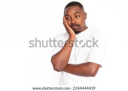 Bored African American man leaning on hand. Portrait of sleepy young male model with short hair and beard in white T-shirt looking away, pouting lips. Boredom, apathy concept
