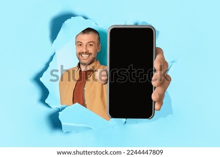 Happy middle aged man holding big smartphone with blank screen in hand, showing gadget with copy space for mockup through torn blue paper hole, collage
