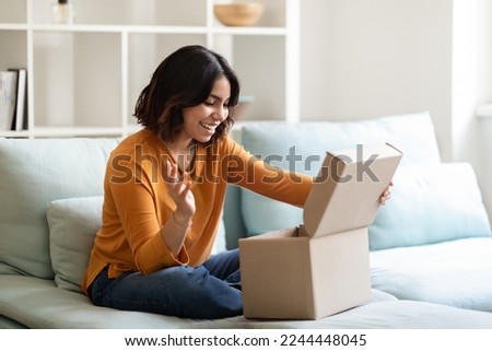 Happy young arab woman opening delivery box at home and looking inside, excited middle eastern lady unboxing parcel with purchases while sitting on couch in cozy living room interior, copy space Royalty-Free Stock Photo #2244448045
