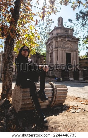 Stylish dark gothic guy between large ancient abandoned mausoleum with neoclassical style with blue sky in a sunny day