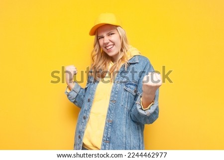 young girl in a denim jacket and a cap rejoice at success on a yellow background, winner girl enjoy victory