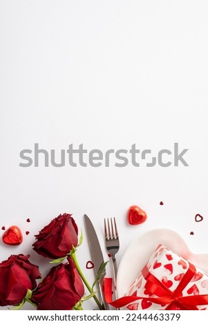 Valentine's Day concept. Top view vertical photo of bouquet of red roses heart shaped plate with giftbox candies knife fork and confetti on isolated white background with empty space Royalty-Free Stock Photo #2244463379
